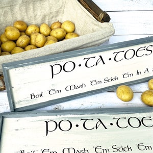 May include: Two rustic wooden signs with white paint and black lettering. The signs read "PO-TA-TOES" and include instructions for preparing potatoes: "Boil 'Em Mash 'Em Stick 'Em In A" and "Boil 'Em Mash 'Em Stick 'Em".