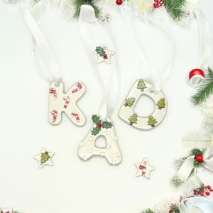 May include: White ceramic Christmas ornaments spelling "KAD" with red and green accents. The "K" features candy cane designs, the "A" has holly, and the "D" has Christmas trees. Each ornament hangs from a white ribbon.