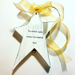 May include: White wooden star ornament with a gold ribbon and a bell. The star has the quote "The darkest nights produce the brightest stars" written on it.