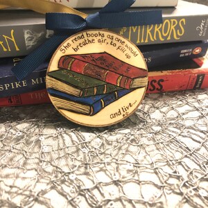May include: A wooden, round ornament featuring a stack of three books in red, green, and blue. The ornament has the quote, "She read books as one would breathe air, to fill up and live..." and is tied with a navy blue ribbon. Books are in the background.