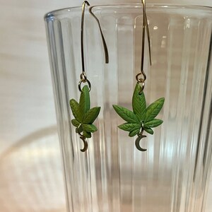 May include: A pair of green cannabis leaf earrings with gold-colored ear wires. Each earring features a detailed leaf design with a small crescent moon charm. The earrings are displayed in front of a clear glass.