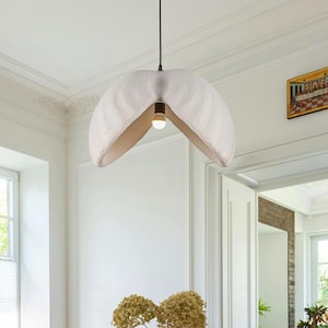 Puede incluir: Una lámpara colgante moderna con una pantalla blanca y un interior dorado cuelga sobre una mesa de comedor redonda. La mesa está puesta con un mantel verde claro y rodeada de sillas de madera. La habitación tiene paredes blancas y suelo de parquet.