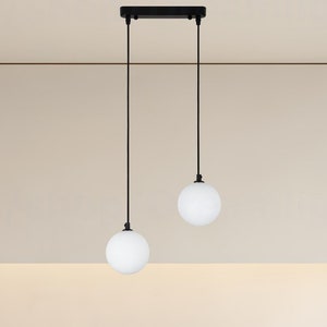 May include: Two white globe pendant lights hang from a black metal track in a modern kitchen. The lights are suspended over a white countertop with a stainless steel sink and a black stove.