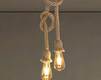 May include: A dual-pendant light fixture featuring two thick, tan-colored twisted hemp ropes tied into decorative knots. Each rope terminates in a flared, frayed rope socket housing a glowing, amber-tinted Edison-style light bulb.