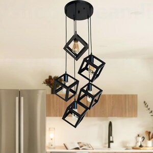 May include: A modern black geometric pendant light fixture with five cube-shaped shades, each housing a lightbulb. The fixture hangs above a wooden dining table with four chairs in a contemporary kitchen setting.