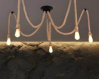 May include: A ceiling light fixture featuring five hanging light bulbs suspended by thick, beige twisted rope cords. The cords are attached to a central black circular mount and extend outward to individual points, creating a draped, spider-like design.