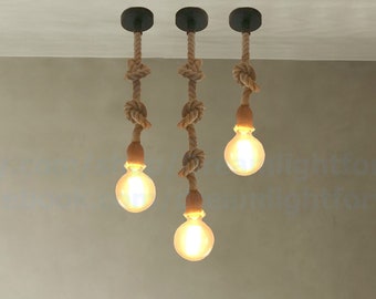 May include: Three pendant lights hanging from the ceiling, each featuring a thick, natural-colored twisted rope cord with decorative knots. Each rope is attached to a black circular canopy and ends in a glowing, round Edison-style light bulb.