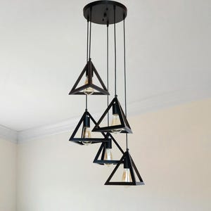 May include: A modern black geometric chandelier with five triangular shades hangs above a wooden dining table. The fixture is suspended from a circular ceiling mount. The room features a neutral color palette with a window and wooden chairs.