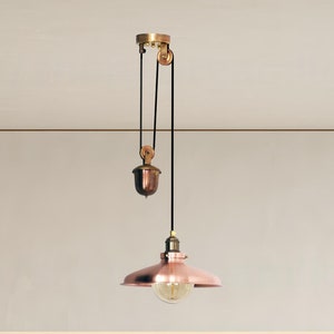 Może przedstawiać: Wisząca lampa miedziana z białym mocowaniem sufitowym i czarnym kablem. Lampa ma miedziany klosz i przezroczystą żarówkę. Lampa wisi nad blatem kuchennym z białym blatem i stalowym zlewozmywakiem.