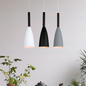 Op de afbeelding: Drie hanglampen met zwarte snoeren en kegelvormige lampenkappen in wit, zwart en grijs, hangend boven een tafel met een vaas met bloemen en andere decoratieve items.