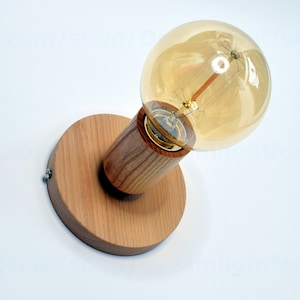 Op de afbeelding: Een houten wandlamp met een ronde basis en een cilindrische steel. Een heldere glazen lamp met een gloeidraad is bevestigd aan de steel.