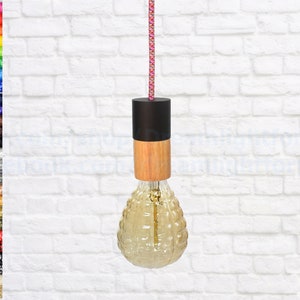 Könnte beinhalten: Eine Pendelleuchte mit einem Holz- und schwarzen Sockel und einer klaren Glasbirne mit strukturierter Oberfläche. Die Leuchte hängt an einem rosa-weißen Kabel vor einer weißen Backsteinwand.