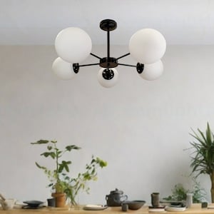 May include: A black metal chandelier with six white globe-shaped light bulbs. The chandelier is hanging from the ceiling in a room with a wooden table and plants.