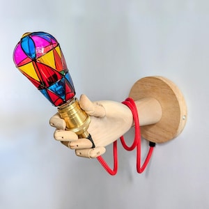 Op de afbeelding: Een houten hand die een kleurrijk glas-in-lood lampje vasthoudt met een rood snoer. De hand is bevestigd aan een houten wandbevestiging.