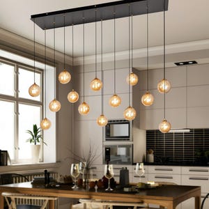 Op de afbeelding: Een moderne eetkamer met een lange houten tafel en stoelen, verlicht door een zwarte rechthoekige kroonluchter met meerdere bolvormige lampen. De lichten zijn amberkleurig en creëren een warme sfeer. De keuken heeft witte kasten en een zwarte betegelde achterwand.