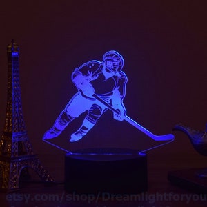May include: A blue LED night light with a silhouette of a hockey player skating with a stick. The light is on a black base and is illuminated with a blue glow.