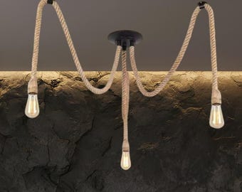May include: A three-light ceiling fixture featuring a central black circular mount with three thick, tan-colored natural fiber ropes extending outward. Each rope ends in a clear glass Edison-style light bulb, creating a hanging pendant design.