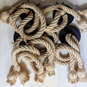 May include: A collection of thick, natural-coloured jute rope segments attached to circular black metal mounting plates. Each rope features frayed, tassel-like ends, creating a textured, industrial appearance with multiple loops and coiled sections.