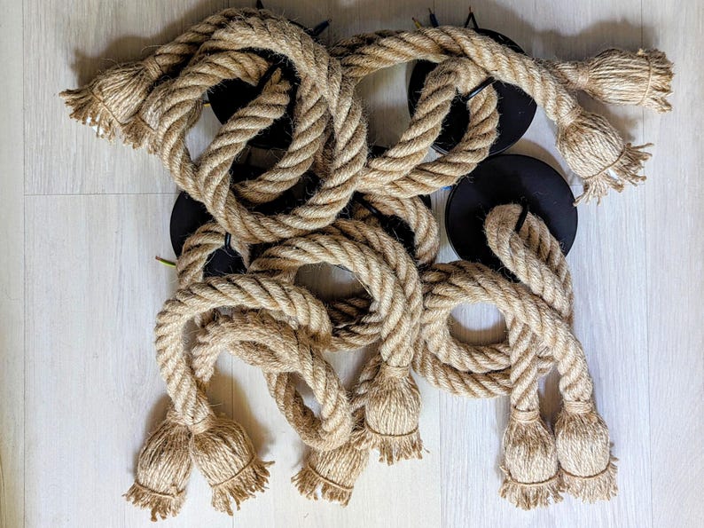 May include: A collection of thick, natural-coloured jute rope segments attached to circular black metal mounting plates. Each rope features frayed, tassel-like ends, creating a textured, industrial appearance with multiple loops and coiled sections.