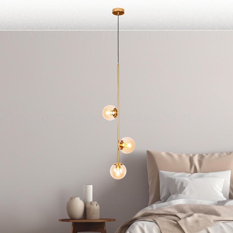 May include: A modern pendant light fixture with three spherical glass shades. The fixture has a gold finish and hangs from a thin black cord. The glass shades are amber-colored, and the light is on. The fixture is above a bed.