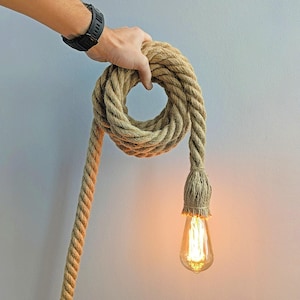 Puede incluir: Una lámpara colgante rústica de cuerda con una bombilla Edison de brillo cálido. La gruesa cuerda de color natural está enrollada y sostenida por una mano, creando una lámpara única y texturizada. La bombilla tiene forma de lágrima.