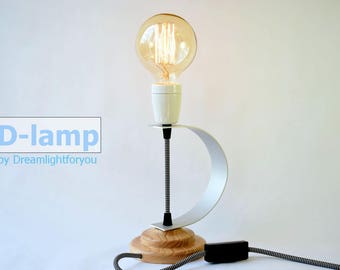 Peut inclure: Une lampe de table blanche et en bois avec un accent en métal argenté et une ampoule de style vintage. La lampe est sur un fond blanc et a un cordon noir.