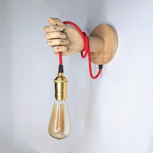 Könnte beinhalten: Eine Holzhand hält ein rotes Kabel mit einer daran befestigten goldenen Glühbirne. Die Hand ist auf einem Holzsockel montiert.