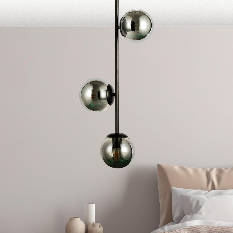 Op de afbeelding: Een moderne hanglamp met drie bolvormige, spiegelende glazen kappen. De kappen hangen aan een zwarte metalen staaf. De lamp hangt in een slaapkamer.
