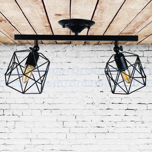 May include: Black, geometric-shaped ceiling light fixture with two bulbs. The fixture has a rectangular bar with two adjustable light heads. The bulbs are visible inside the geometric cages. The background is a white brick wall and wooden ceiling.