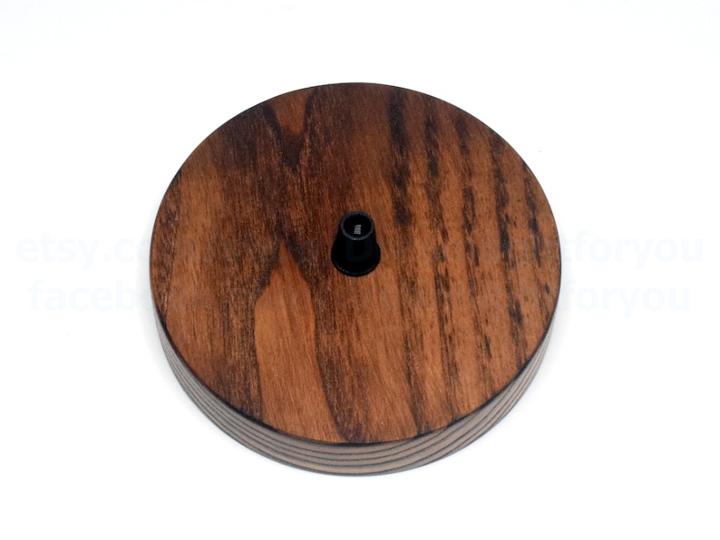 K&ouml;nnte beinhalten: Eine runde Deckenrosette aus Holz mit einer dunkelbraunen Beize. Die Rosette hat eine schwarze elektrische Armatur in der Mitte.