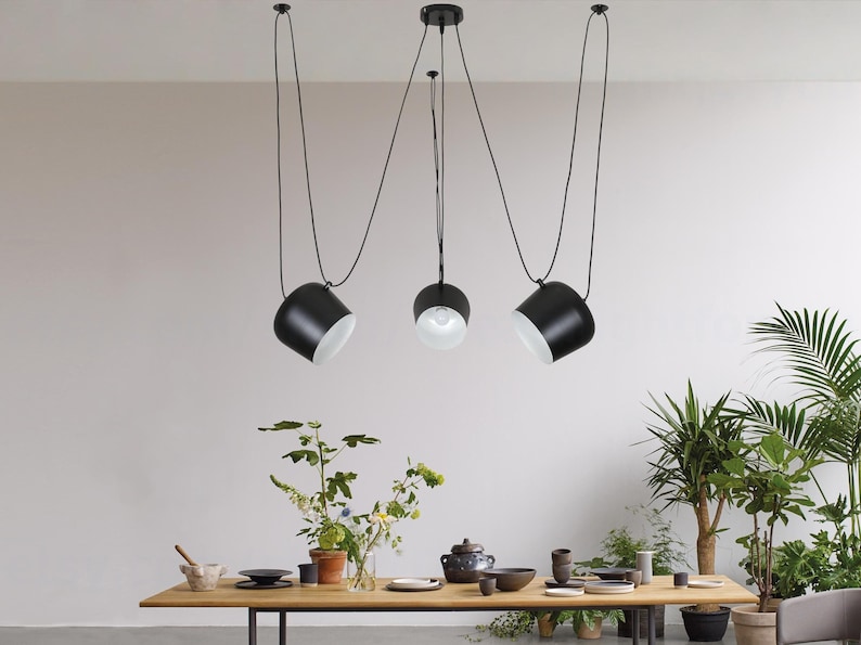 May include: Three black pendant lights with white interior shades hang from the ceiling over a wooden table with plants and dishes on it.