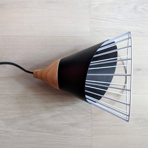 Pu&ograve; includere: Una lampada a sospensione con un paralume conico in metallo nero fissato a una base in legno naturale. Il paralume presenta un'estensione a gabbia in filo metallico bianco sull'apertura. Un cavo elettrico nero esce dalla parte superiore in legno.