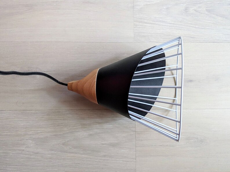 Pu&ograve; includere: Una lampada a sospensione con un paralume conico in metallo nero fissato a una base in legno naturale. Il paralume presenta un'estensione a gabbia in filo metallico bianco sull'apertura. Un cavo elettrico nero esce dalla parte superiore in legno.