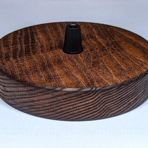K&ouml;nnte beinhalten: Eine runde Deckenrosette aus Holz mit einer dunkelbraunen Beize. Die Rosette hat eine schwarze elektrische Armatur in der Mitte.