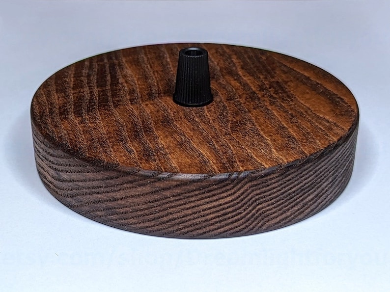 K&ouml;nnte beinhalten: Eine runde Deckenrosette aus Holz mit einer dunkelbraunen Beize. Die Rosette hat eine schwarze elektrische Armatur in der Mitte.