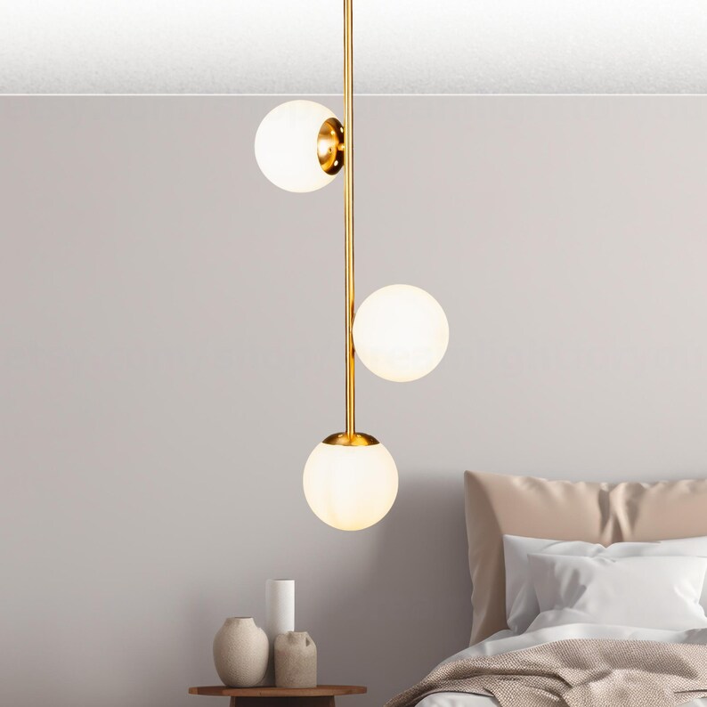 Op de afbeelding: Een moderne hanglamp met drie bolvormige, matglazen kappen. De armatuur heeft een goudkleurig metalen frame en hangt verticaal. Het licht hangt boven een bed met een neutrale kleurstelling.