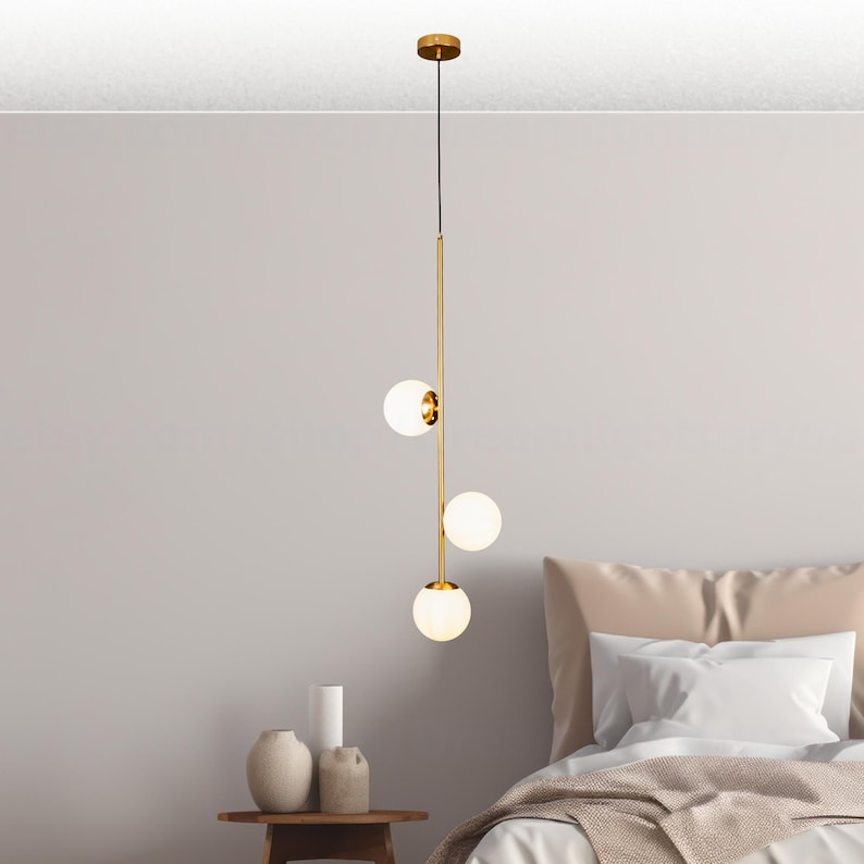 Op de afbeelding: Een moderne hanglamp met drie bolvormige, matglazen kappen. De armatuur heeft een gouden afwerking en hangt aan een dun zwart snoer. Het licht hangt boven een bed.