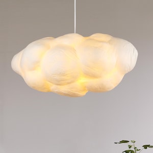 May include: A cloud-shaped pendant light fixture. The light is a soft white color, with a textured surface. The fixture is suspended from a white cord, creating a whimsical and dreamy aesthetic.