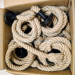 May include: A cardboard box containing several coiled lengths of thick, natural-coloured jute rope. Each rope segment is attached to a circular, matte black metal wall mount fixture. The ropes are arranged in a circular pattern within the box.