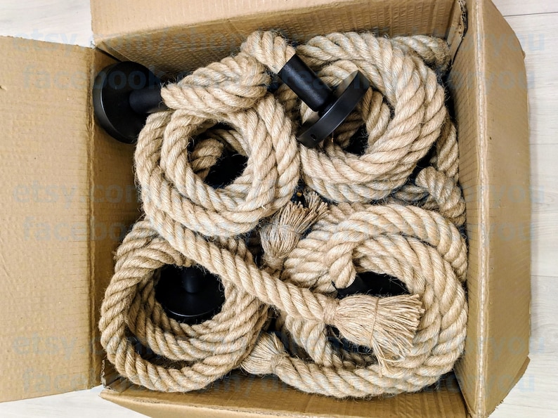 May include: A cardboard box containing several coiled lengths of thick, natural-coloured jute rope. Each rope segment is attached to a circular, matte black metal wall mount fixture. The ropes are arranged in a circular pattern within the box.