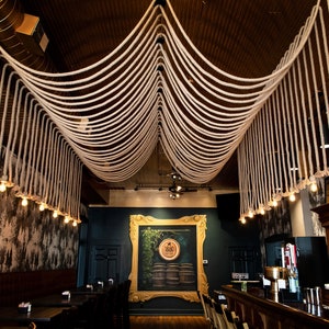 May include: A ceiling installation featuring thick, off-white twisted ropes draped in symmetrical loops across the room, with small, warm-toned Edison-style light bulbs hanging from the ends of the ropes, creating a decorative lighting display.