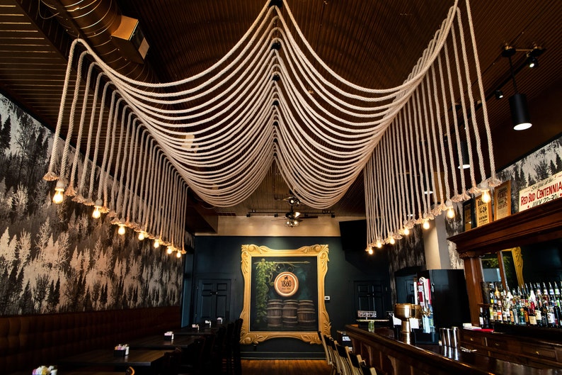 May include: A ceiling installation featuring thick, off-white twisted ropes draped in symmetrical loops across the room, with small, warm-toned Edison-style light bulbs hanging from the ends of the ropes, creating a decorative lighting display.