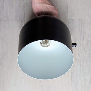 May include: A black and white pendant light fixture with a white interior and a black exterior. The light is turned on and the bulb is visible.