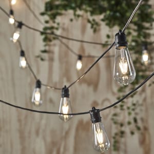 May include: A string of clear, teardrop-shaped light bulbs on a black wire. The bulbs are illuminated, giving off a warm light. The background shows a wooden surface and green leaves, creating a cosy atmosphere. Ideal for indoor or outdoor decoration.