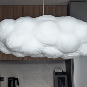 May include: A large, white, cloud-shaped pendant light fixture with a textured, rounded surface. The lamp is suspended from the ceiling by a thin white cord, featuring a voluminous, multi-lobed form that mimics the appearance of a cumulus cloud.