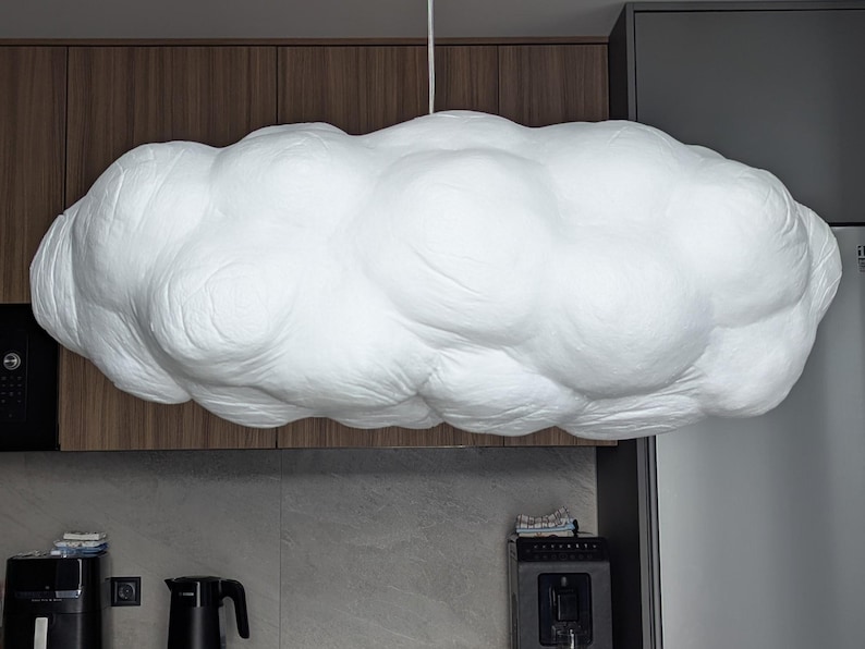 May include: A large, white, cloud-shaped pendant light fixture with a textured, rounded surface. The lamp is suspended from the ceiling by a thin white cord, featuring a voluminous, multi-lobed form that mimics the appearance of a cumulus cloud.