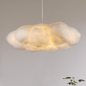 May include: A cloud-shaped pendant light fixture. The light is white and fluffy, resembling a cumulus cloud. The light is illuminated from within, creating a soft, warm glow. The fixture hangs from a white cord.