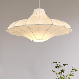 May include: A white, scalloped-edge pendant light fixture. The light has a rounded, inverted-bowl shape with vertical seams and a soft glow. The fixture is suspended from a white cord, suitable for interior lighting.