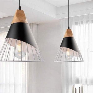 Pu&ograve; includere: Due lampade a sospensione con una sezione superiore conica in legno chiaro, una fascia centrale nera opaca e una base a gabbia metallica bianca. Ogni lampada pende da un cavo nero e contiene una lampadina visibile all'interno.