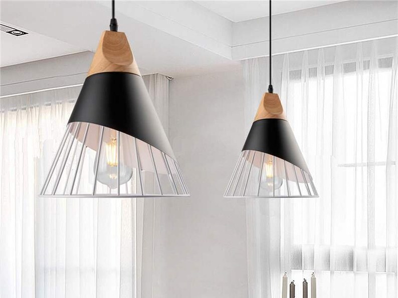 Pu&ograve; includere: Due lampade a sospensione con una sezione superiore conica in legno chiaro, una fascia centrale nera opaca e una base a gabbia metallica bianca. Ogni lampada pende da un cavo nero e contiene una lampadina visibile all'interno.